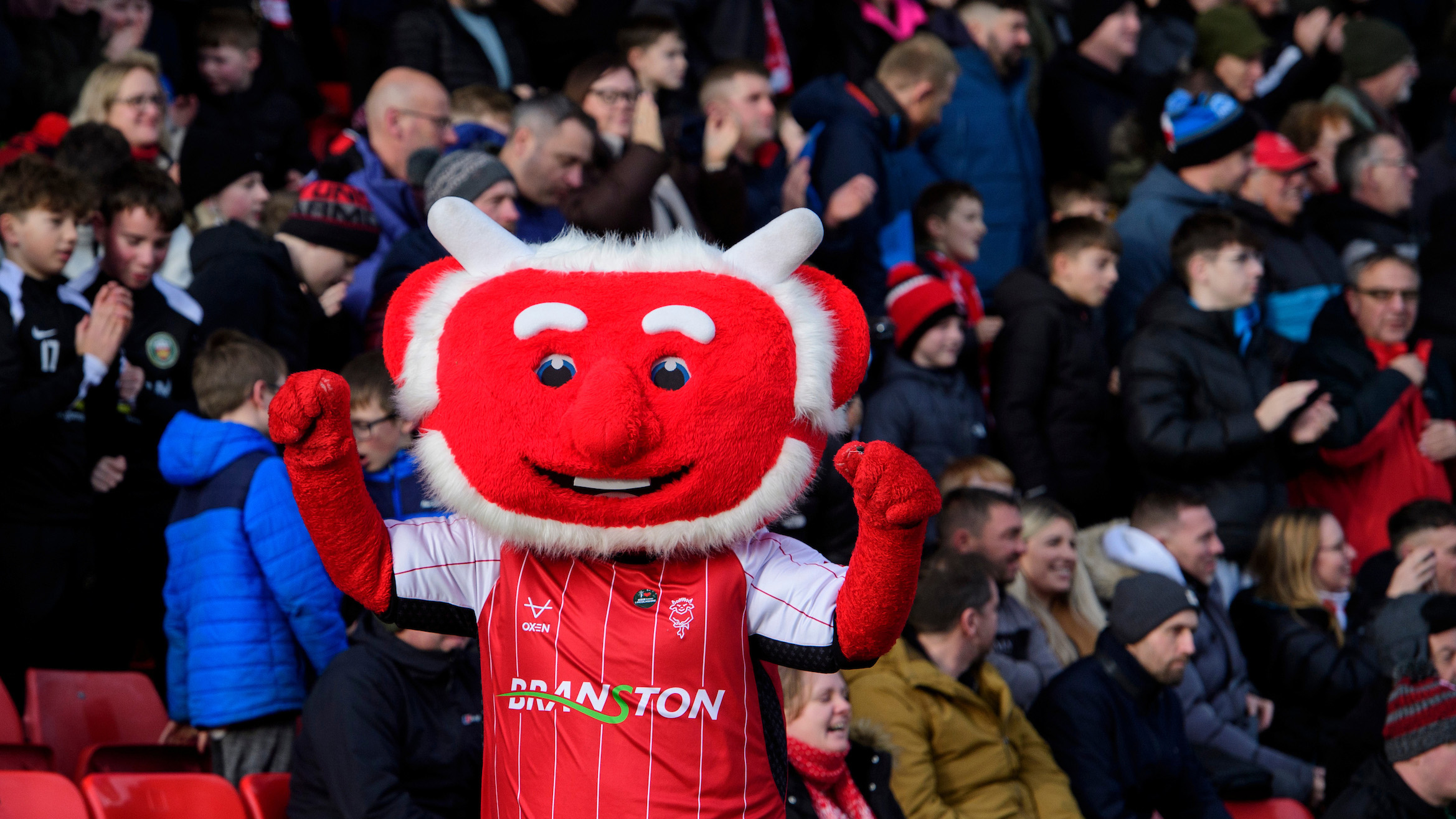 Poacher the Imp celebrates in front of supporters. Poacher, who has a large red head, is wearing a red home Lincoln City shirt with white sleeves. He is holding both arms up in celebration.