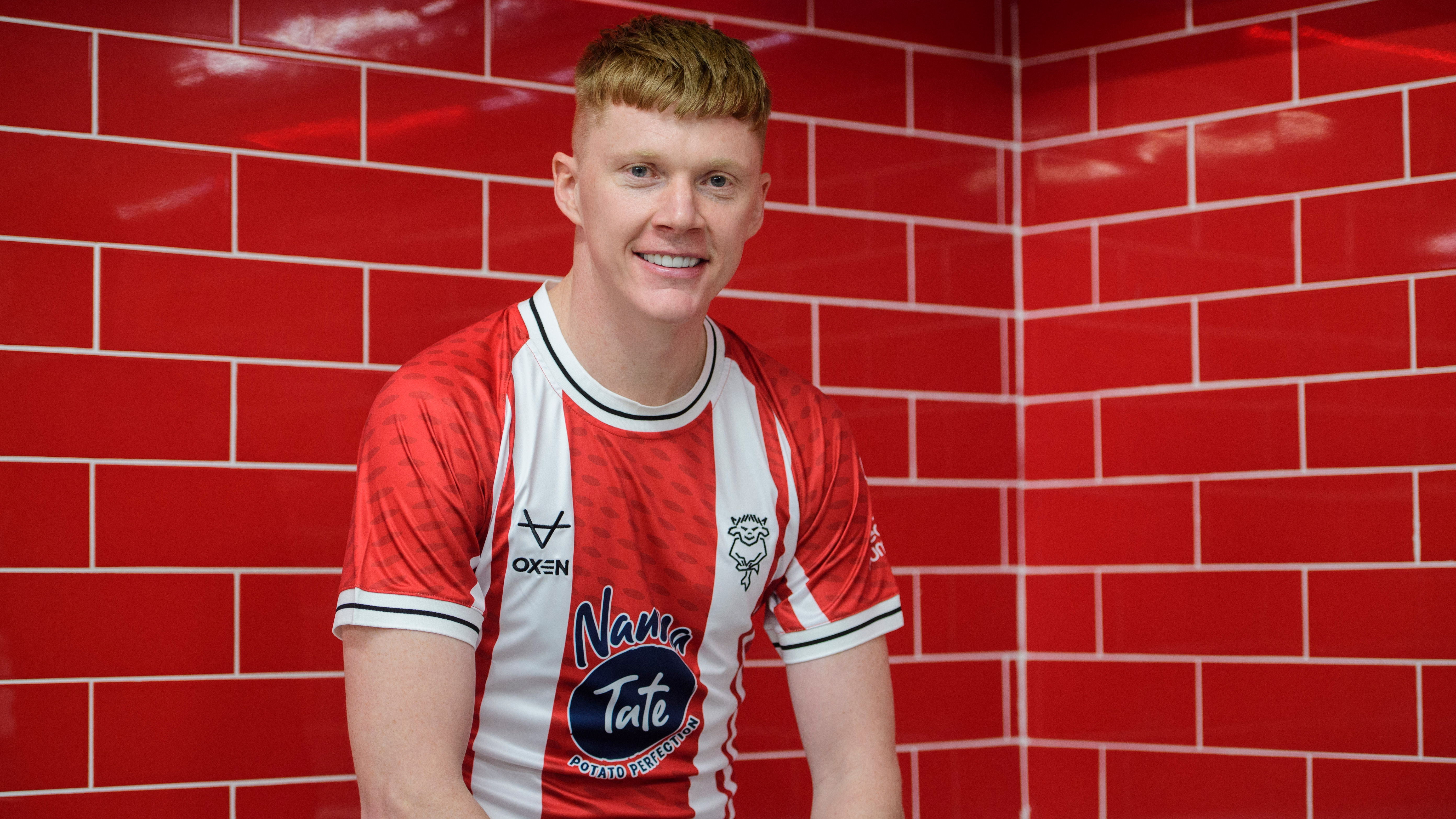 Sam Clucas sits in a changing room. He is wearing a red and white striped shirt and the background behind him is red tiles.