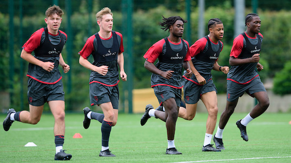 Five footballers running on a training pitch. They are wearing red and grey training kit.