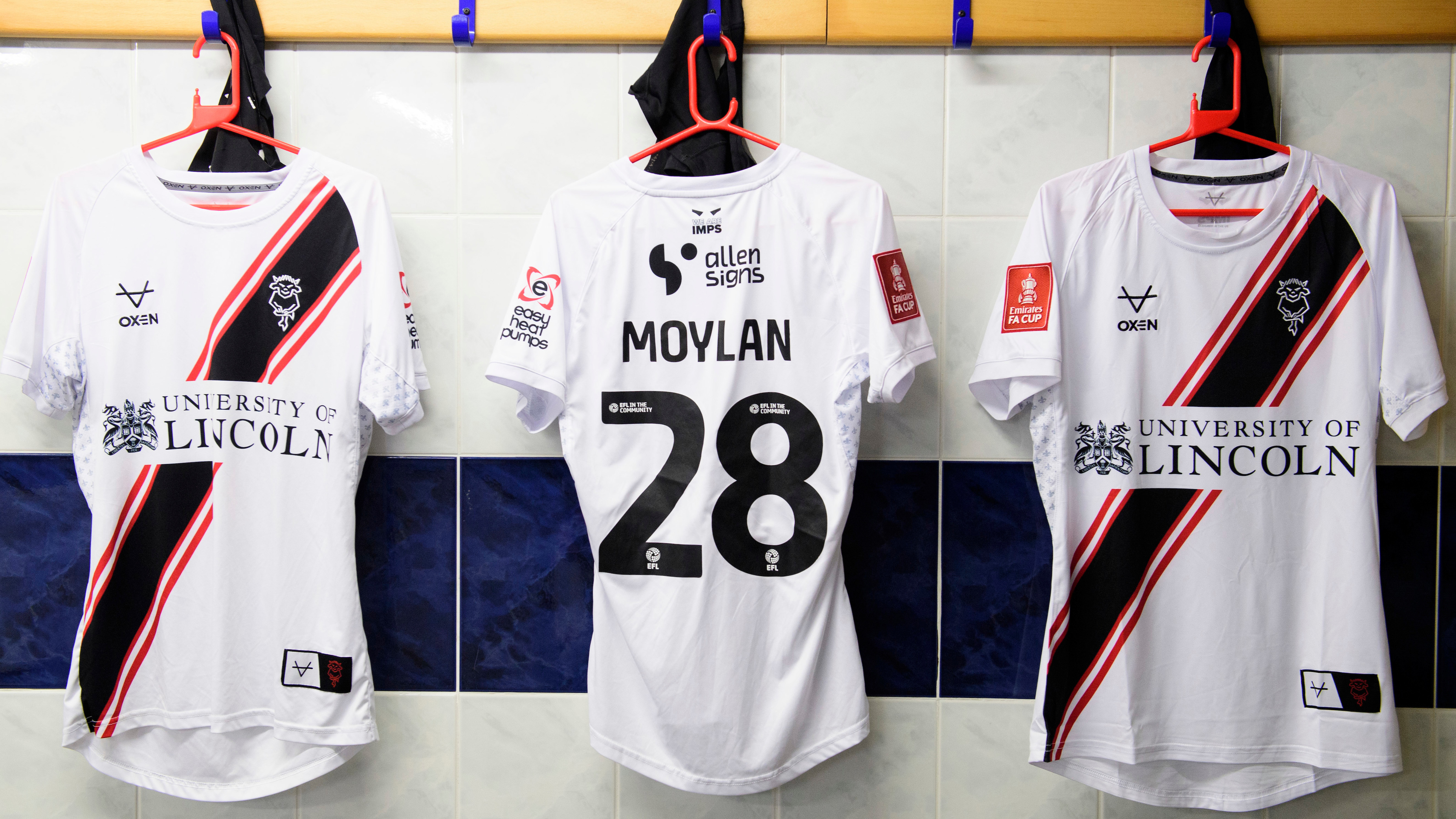 Three white football shirts hung up on pegs in a changing room. The middle shirt has "Moylan 28" on it. The two outside shirts have a sponsor visible which reads "University of Lincoln"