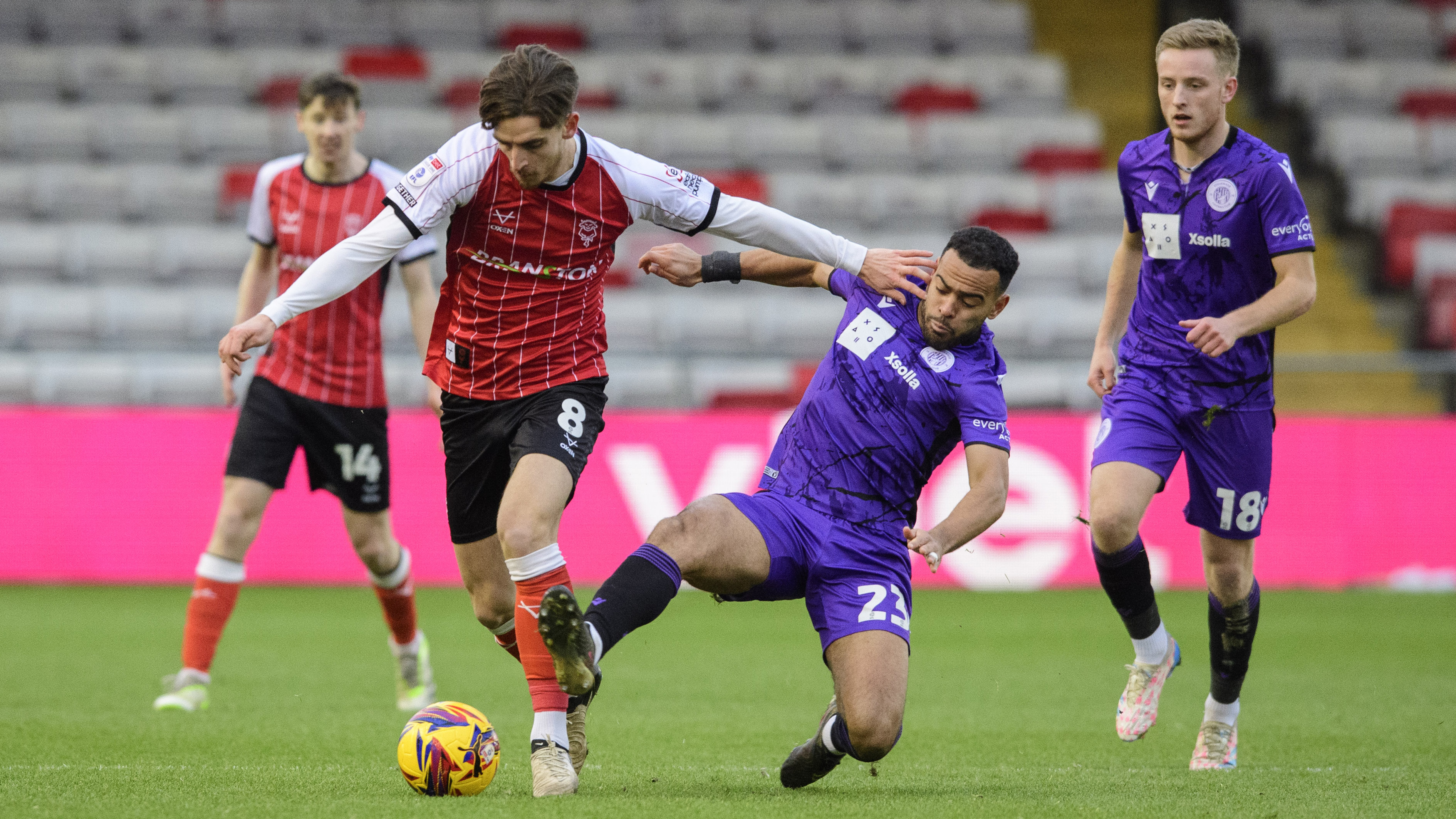 Tom Bayliss, wearing a red shirt and black shorts, tries to get away from a Stevenage player in an all purple kit.