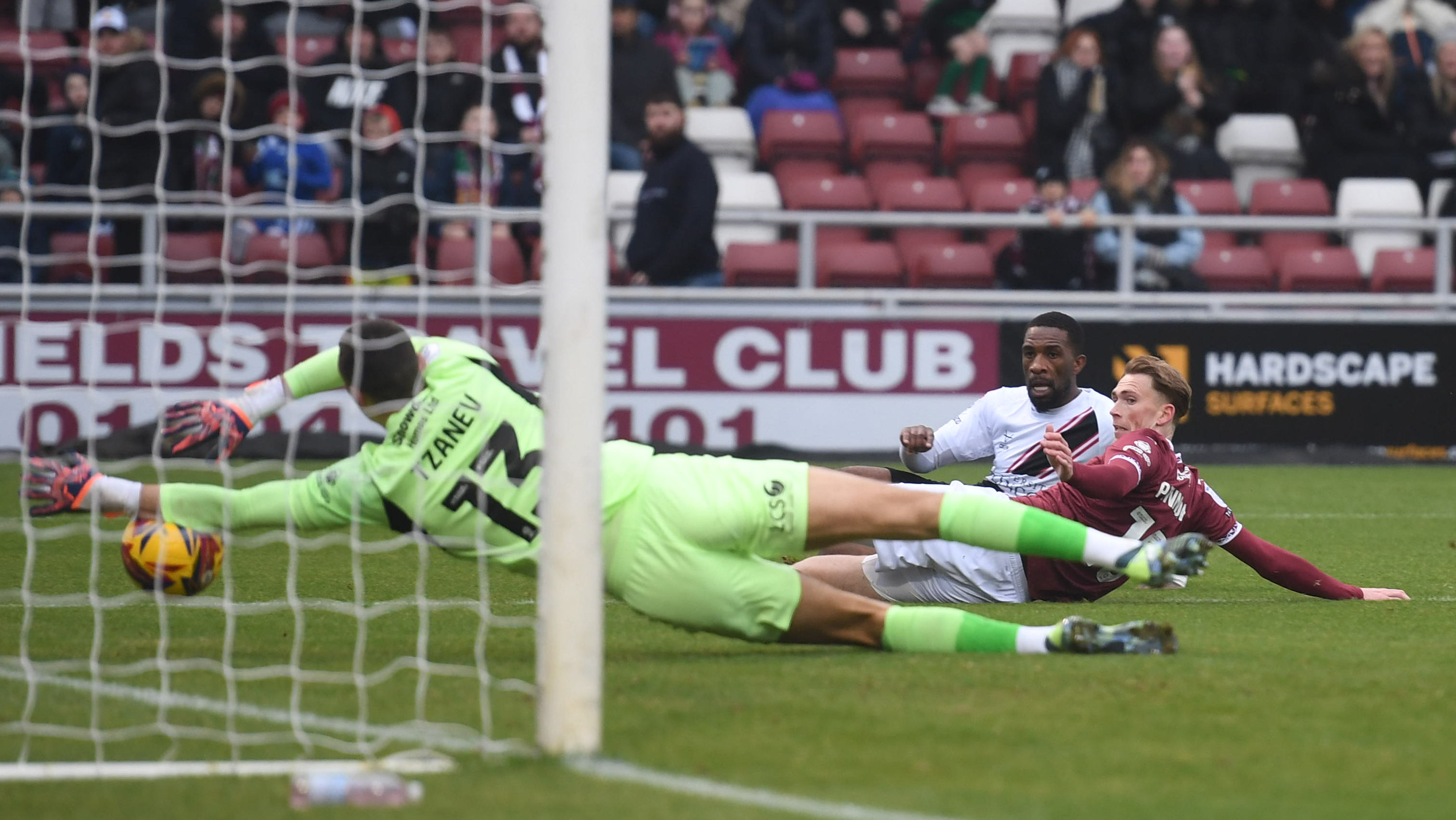 Tendayi Darikwa scores a goal for Lincoln City. A goalkeeper, wearing green, is outstretched trying to get his left hand to the ball as it heads into the net. Behind them Tendayi can be seen watching the ball go over the line.