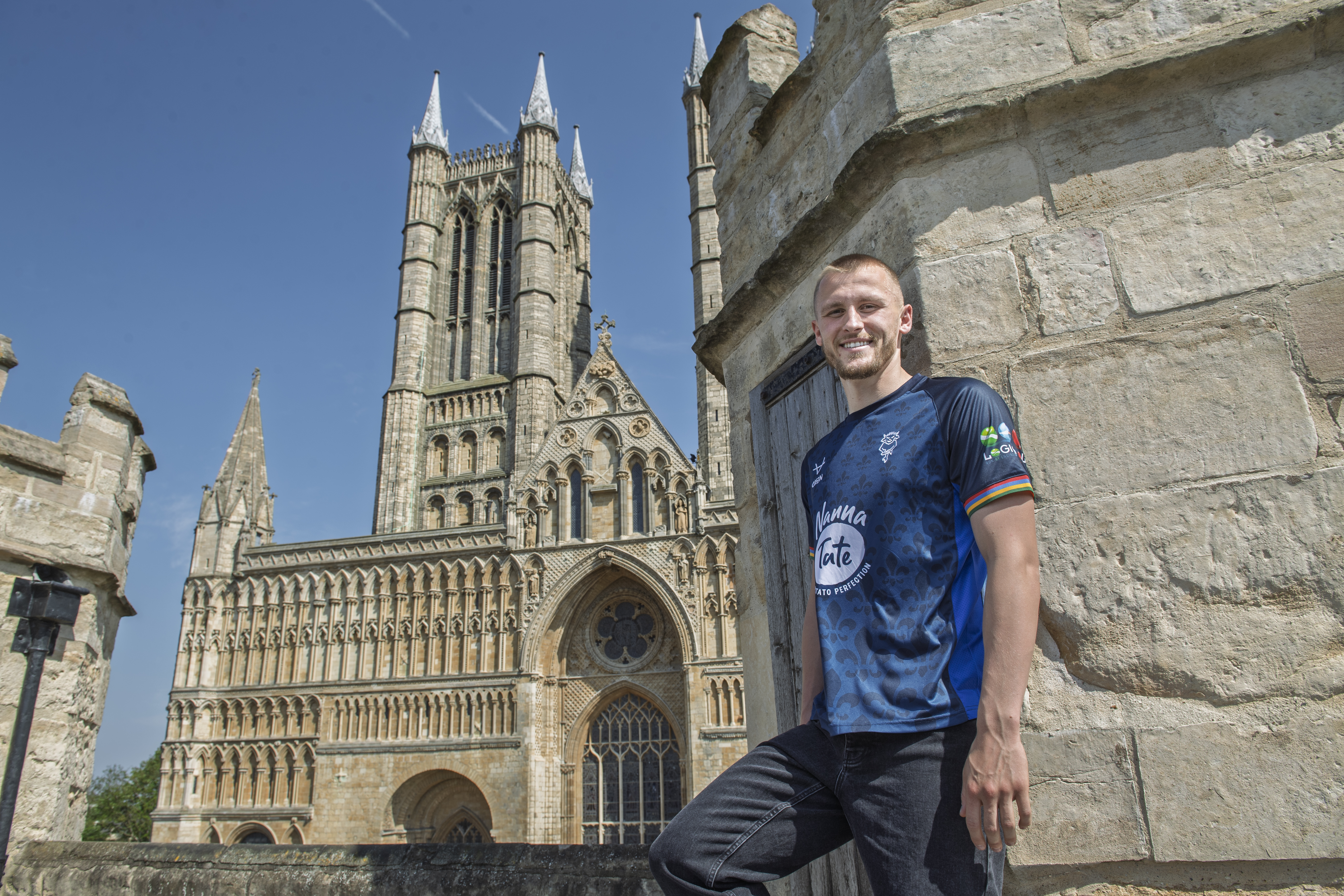 Tom Hamer stands wearing a blue Lincoln City shirt in front of the Western front of Lincoln Cathedral