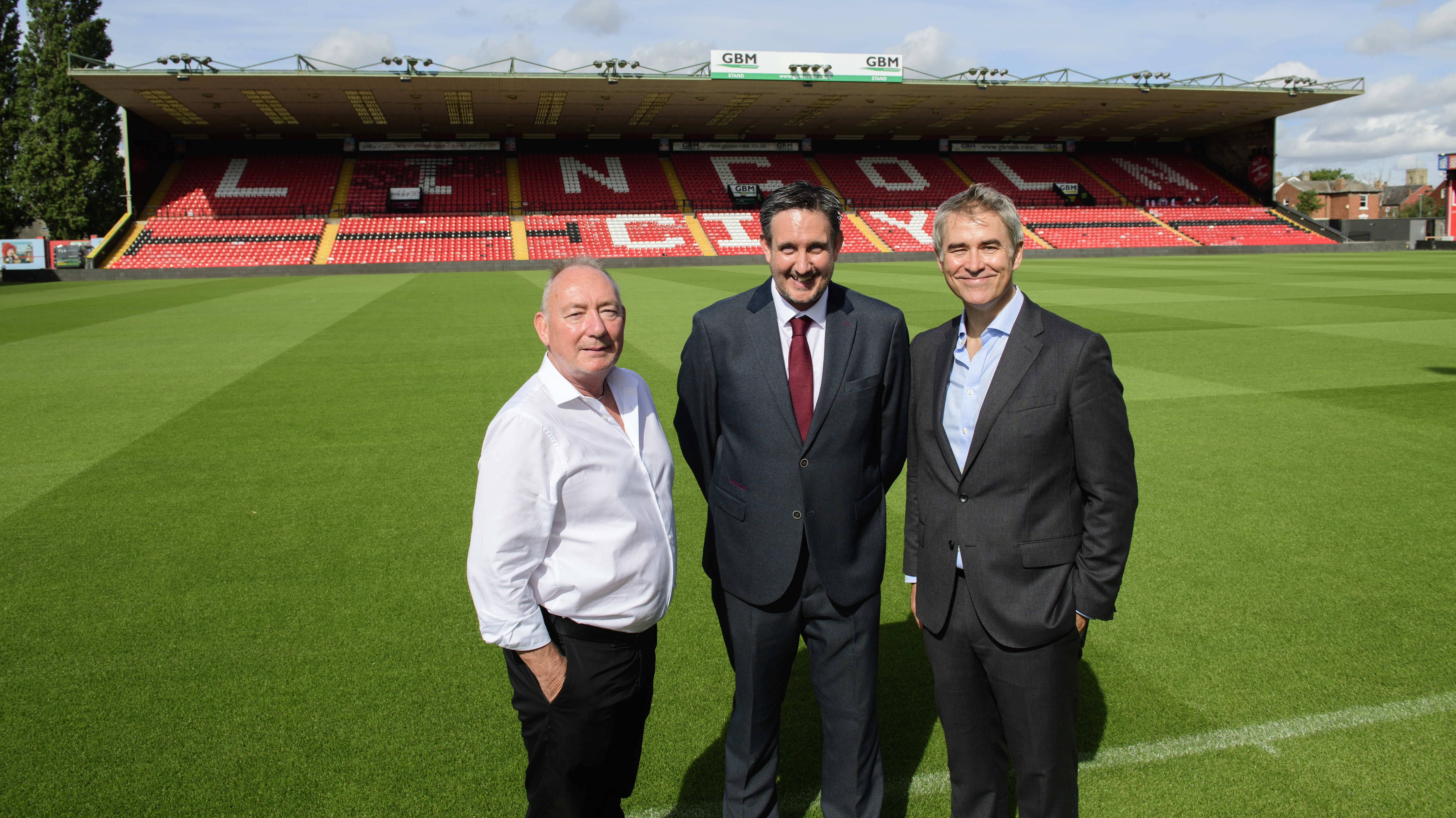 Three men stand in front of a football pitch with an empty stand in the background