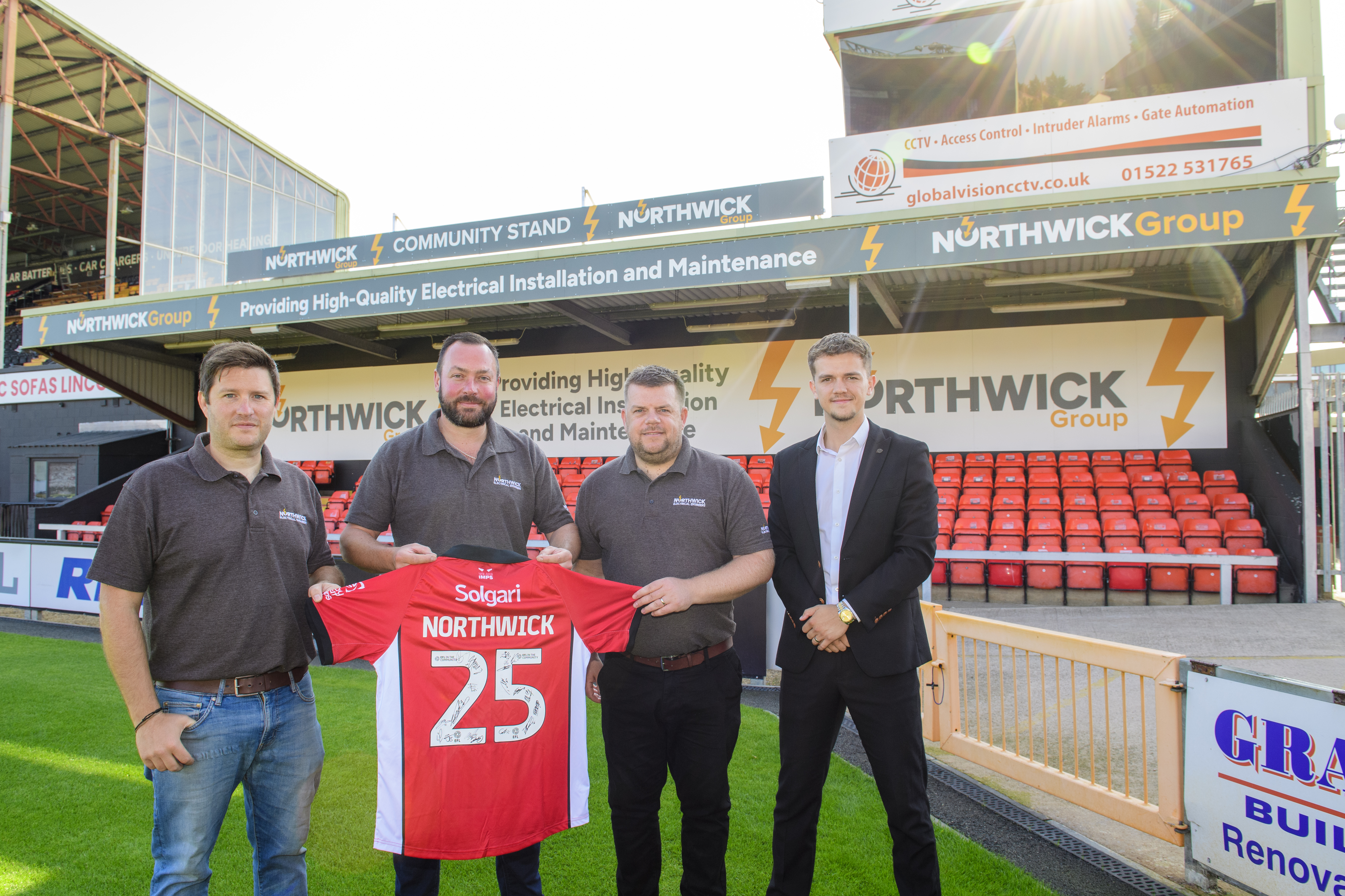 Four people stand holding a red shirt with Northwick 25 on the back