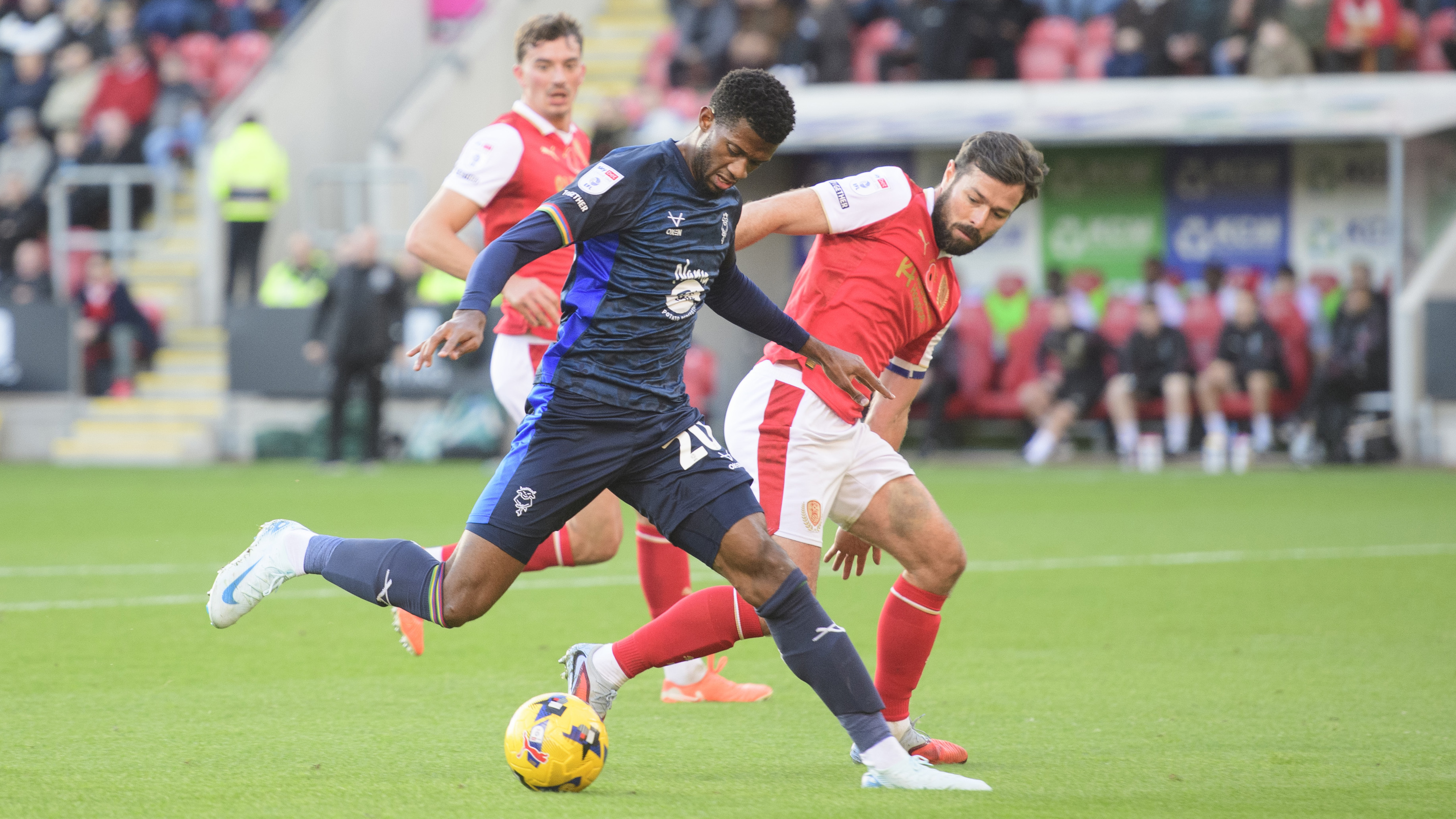 Justin Obikwu, wearing blue t-shirt and shorts, controls the football under pressure from a Rotherham United defender