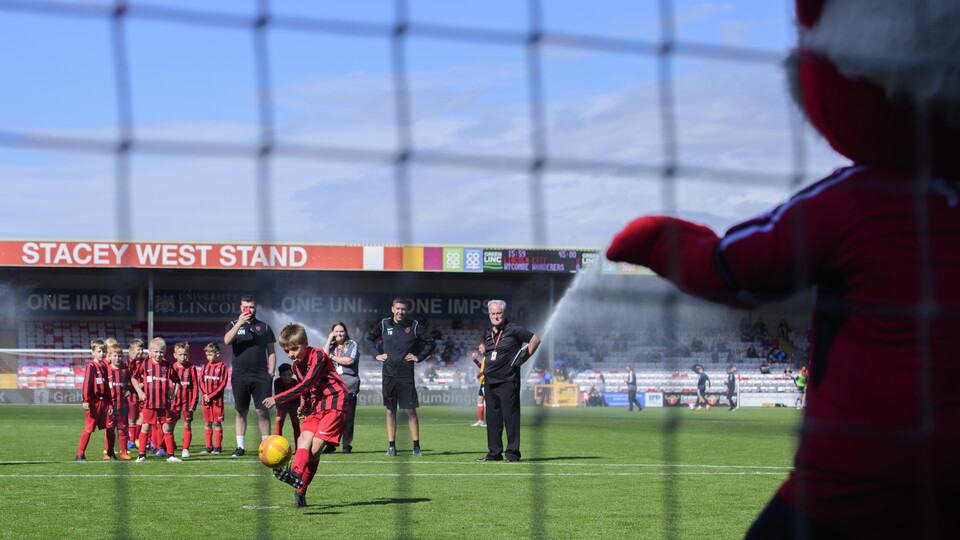 A young boy in a red football kit takes a penalty against Poacher while on the pitch at the LNER Stadium. He scores the goal.