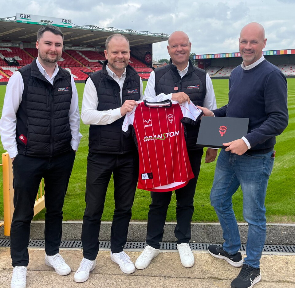 Four men stand at the side of the pitch at the LNER Stadium, holding a 24/25 home shirt and a black box with the Lincoln City logo on.