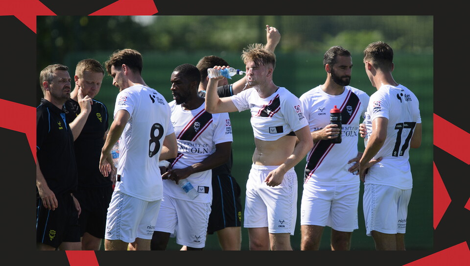 Lincoln City players rehydrate during a water break. JJ McKiernan, in the middle of the group, is shown squirting water into his face from a bottle.