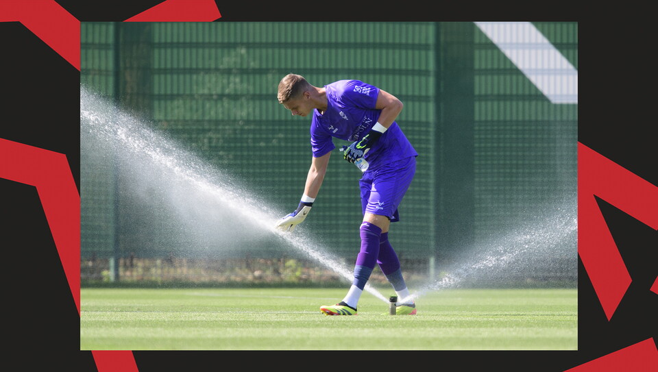 Lincoln City goalkeeper George Wickens, wearing all purple, puts his right hand into a stream of water flowing from a sprinkler