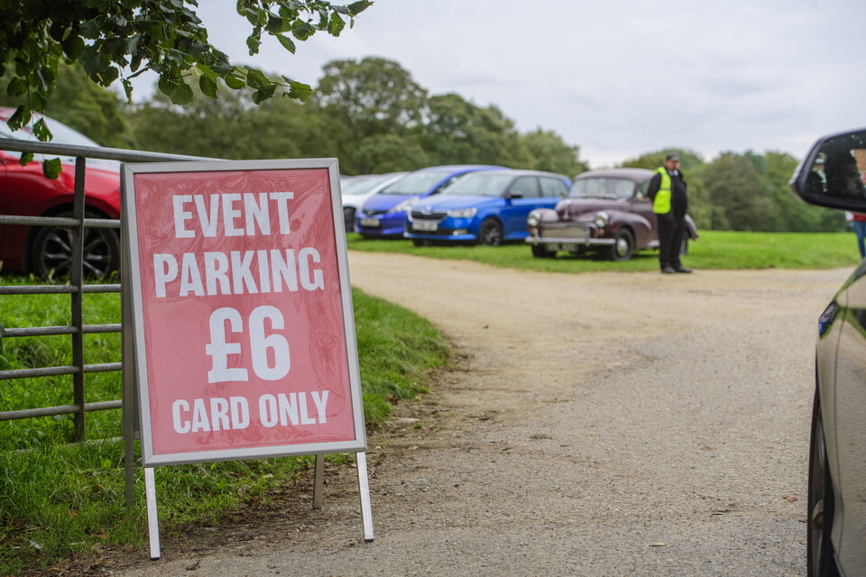 A red sign with white writing on which reads: "Event parking £6 card only" at the entrance to a car park. There are several cars parked in a line in the background.