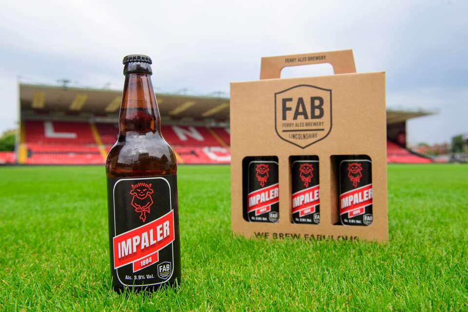 A bottle of Impaler 1884 beer and then a pack of three inside a presentation set, on the green grass of the pitch at the LNER Stadium. The GMB Stand can seen in the background.