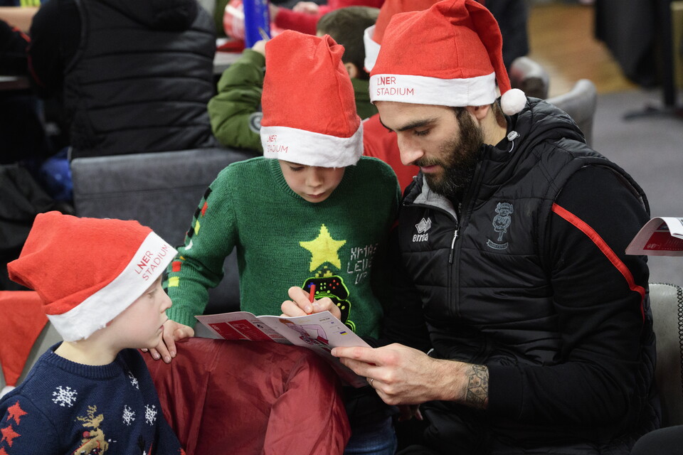 Footballer Adam Jackson looks at a programme along with two children. All three are wearing Santa hats.