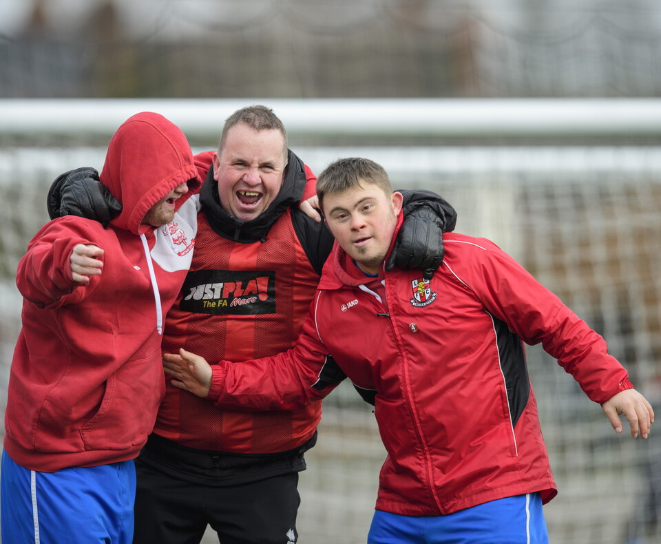 A football coach stands of the middle of two players. He is hugging them in celebration. All three are wearing red tops.