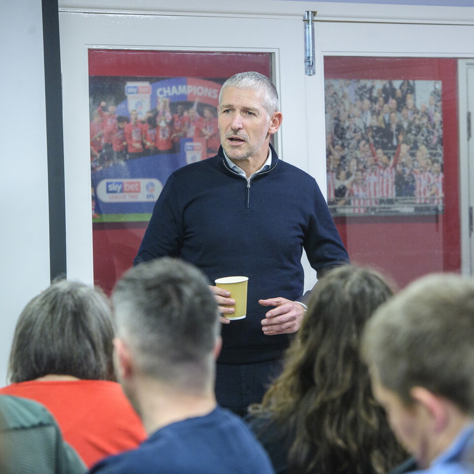 Jez George stands in front a group of people. He is wearing a blue top and holding a drink in his right hand.