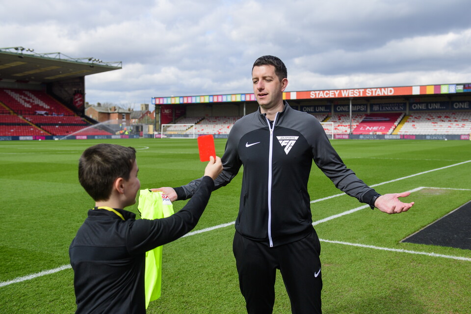 A child holds up his right hand, which is holding a red card, to a referee