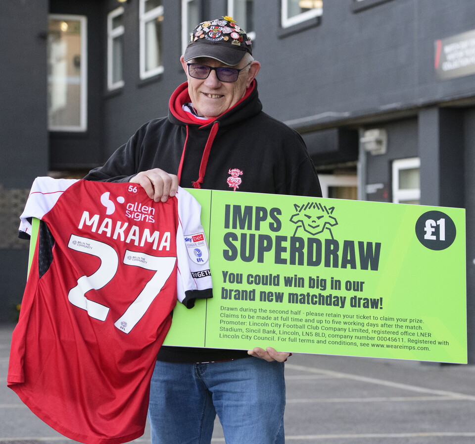 A man holds a green sign with "IMPS SUPERDRAW" written on it. He is holding a shirt with "MAKAMA 27" written on it.
