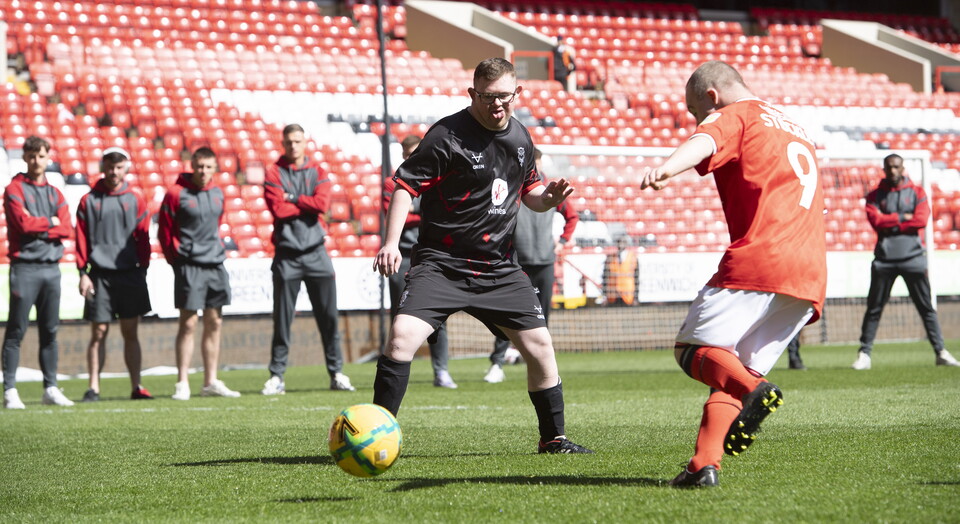 Two people play football while a number of other people stand behind them watching