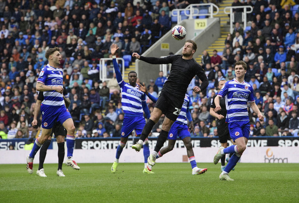 James Collins is mid air having just headed the ball towards goal to put Lincoln City 1-0 up. There are four Reading defenders in blue and white hooped shirts behind him.