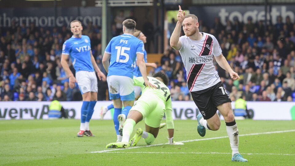 Ben House, wearing a white shirt and black shorts, holds his right hand up in celebration after scoring.