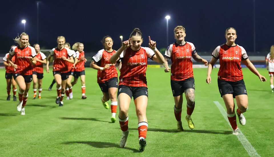 Lincoln City Women players celebrate a goal. They are wearing red shirts and black shorts, and running towards the photographer.