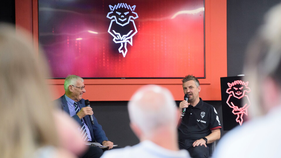 John Helm and Michael Skubala speak on stage. Int he foreground there are people watching.