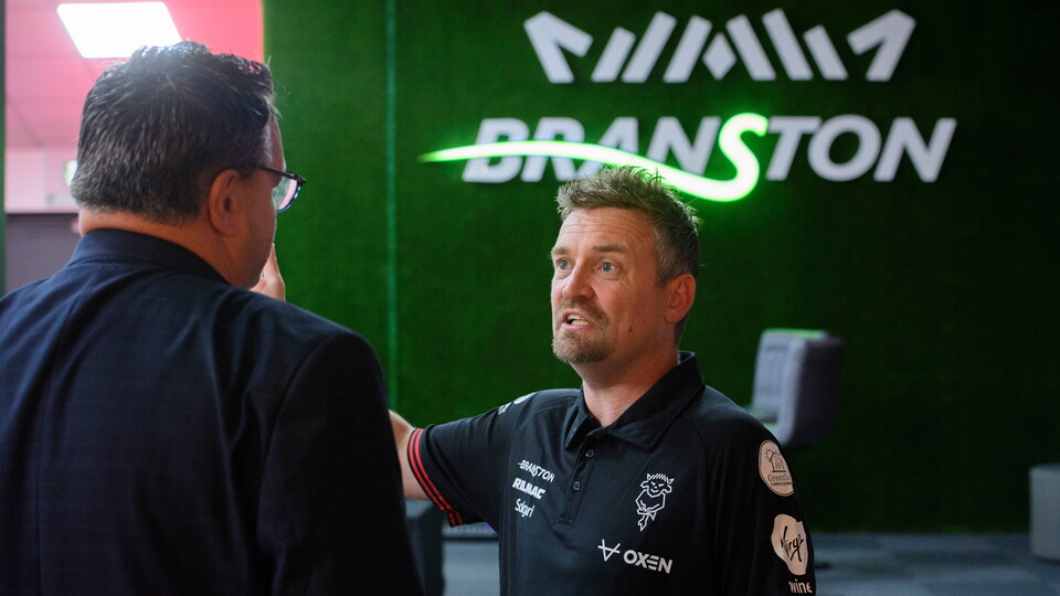 Two men talk in front of a sign which reads Branston.