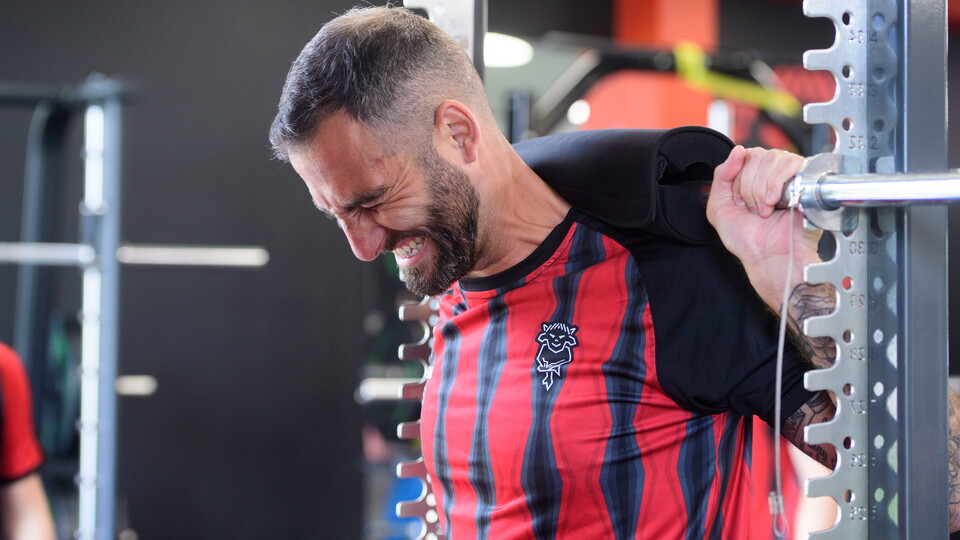 A man grimaces as he pushes his back against a bar in a gym.