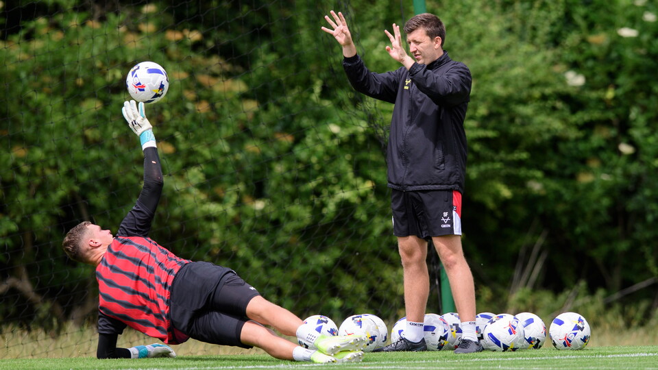 A goalkeeper lays on the ground but holds his right hand up to catch a ball through by a man wearing a black jacket.
