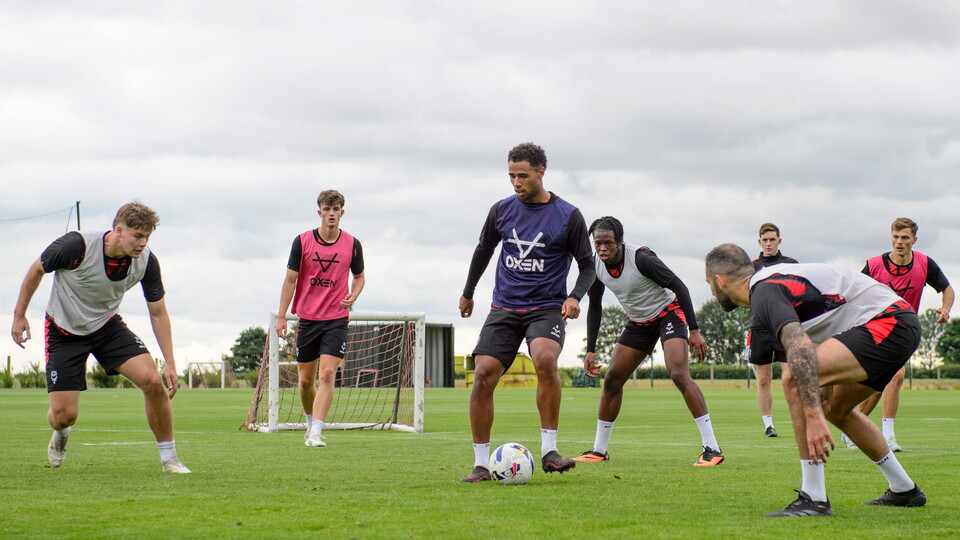 A group of men take part in a game of football. They are on a grass pitch.