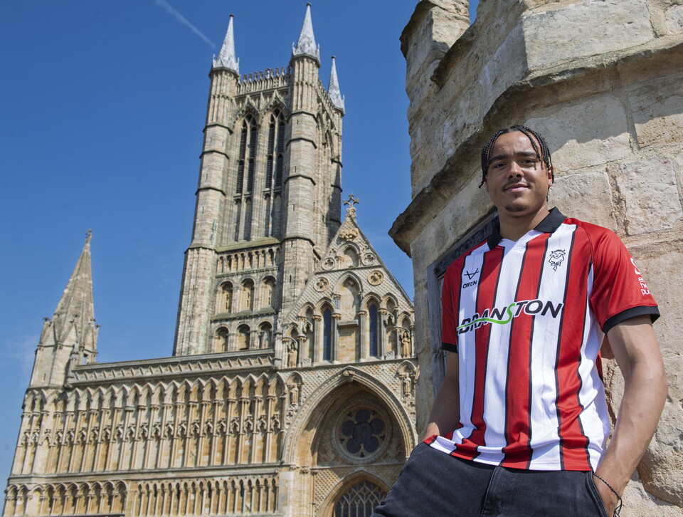 A man stands in a red and white t-shirt, he is leaning against a wall. In the background is the western front of Lincoln Cathedtral.