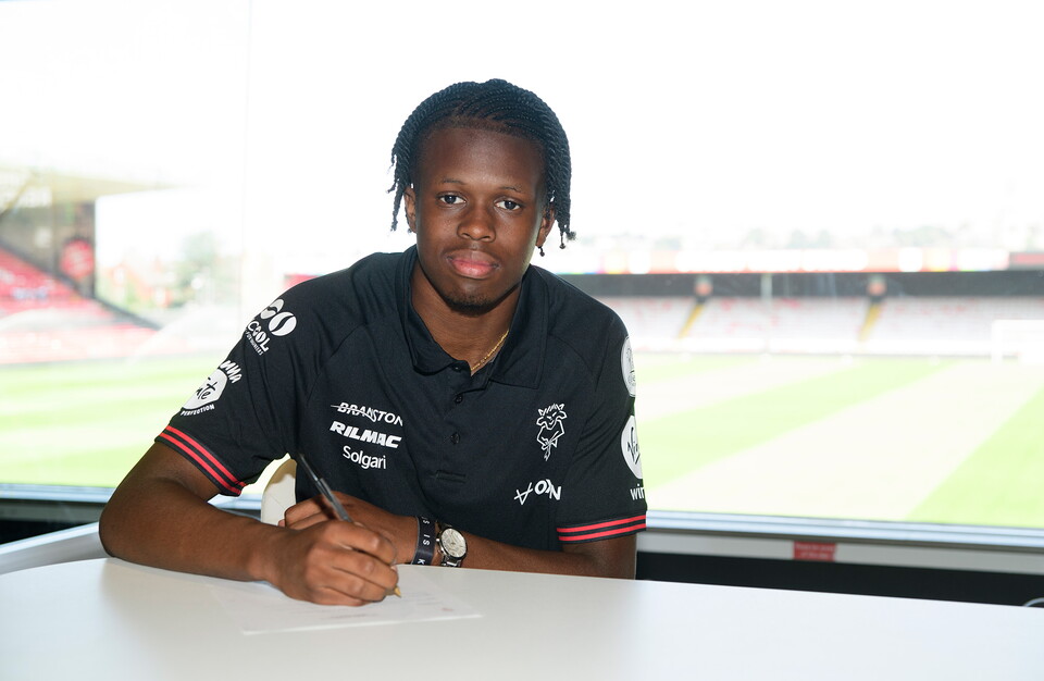 Zane Okoro holds a pen with his right hand to sign a contract. He is wearing a black polo shirt.