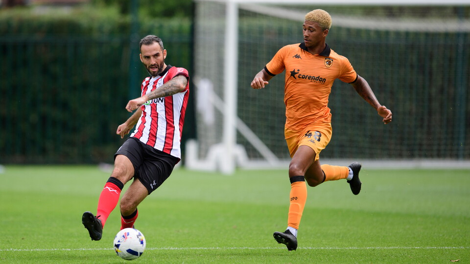 Adam Jackson kicks a ball forward with his right foot. He is chased by another player.