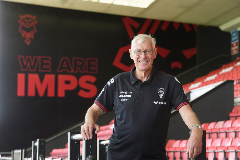 Broadcaster John Helm stands in front of a mural which reads We Are Imps. He is wearing a black polo.