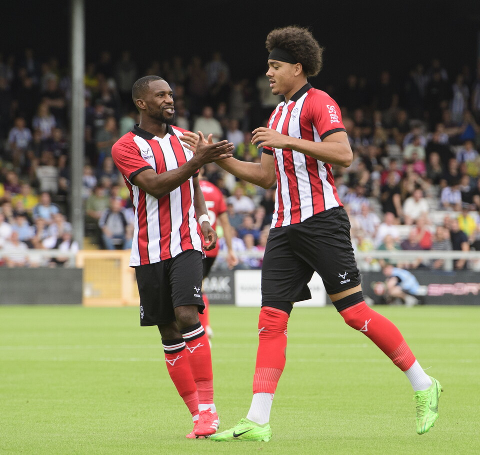 Tendayi Darikwa (left) and Jovon Makama (right) celebrate a goal. They are holding their right hands together. Both are wearing red and white striped tops, black shorts and red socks.