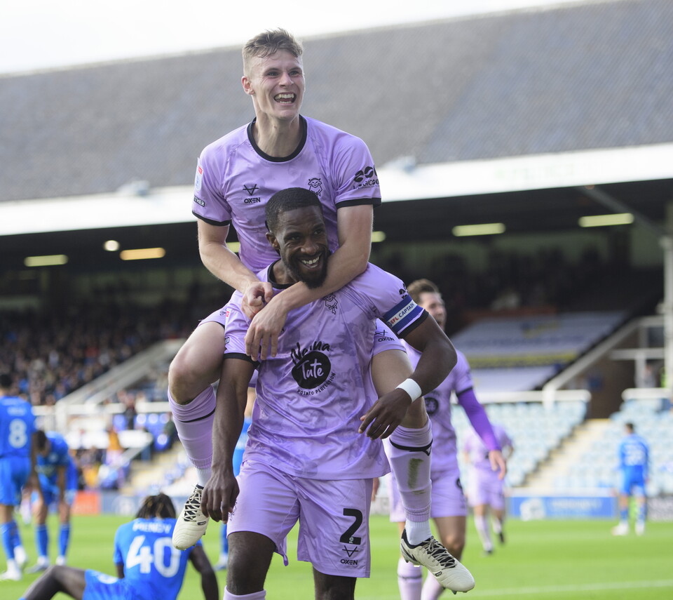 Two footballers in violet kits celebrate a goal. One man is jumping onto the back of the second.