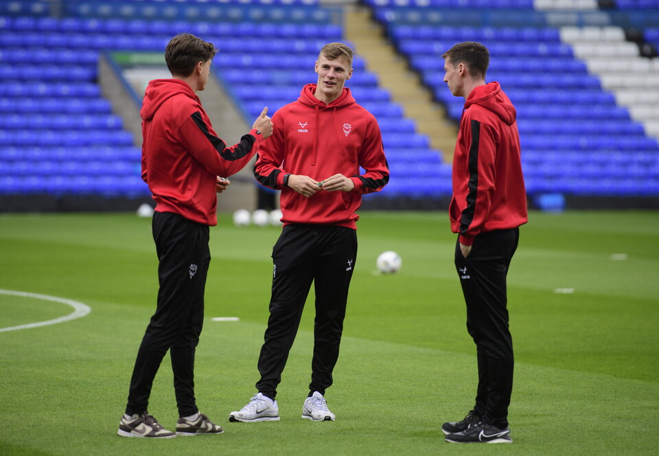 Three men in red hoodies and black tracksuit bottoms stand on a football pitch. In the background is a stand with blue seats.