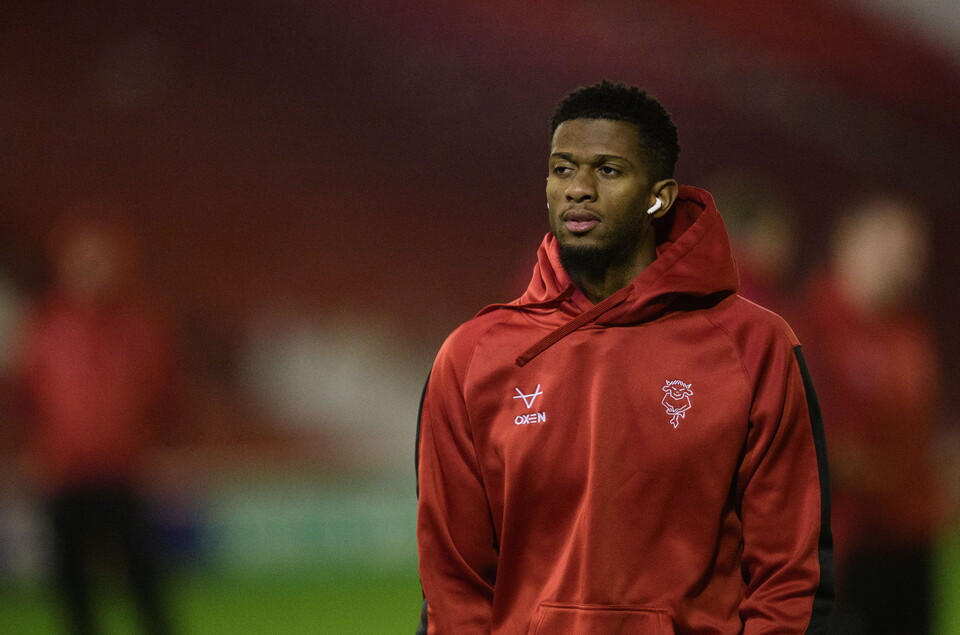 Justin Obikwu stands looking to the left of the image. He is wearing a red hoody.