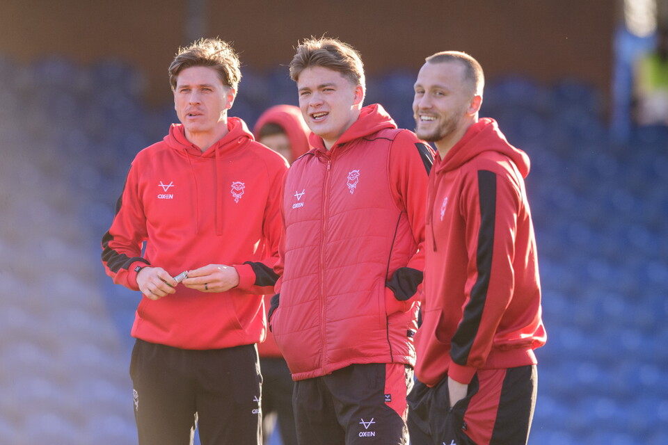 Lincoln City players Adam Reach, Freddie Draper and Tom Hamer stand and look at something off screen. They are wearing red hoodies and black shorts. In the background are blue football stand seats.