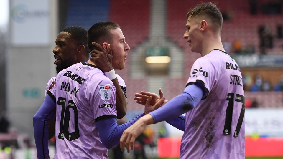 Three players in lilac shirts celebrate a goal in a stadium with blue and red seats