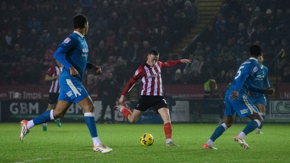 A football player in a red and white striped kit prepares to kick the ball amid opponents in blue kits.