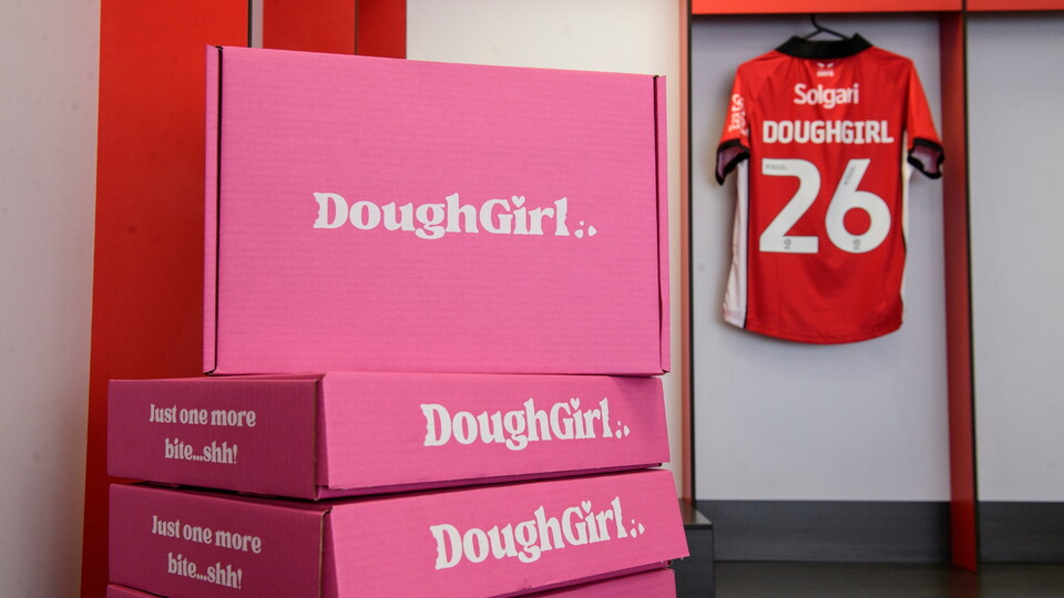 A stack of pink DoughGirl boxes in a changing room. In the background, a Lincoln City football shirt with 'DOUGHGIRL 26' hangs on the wall.