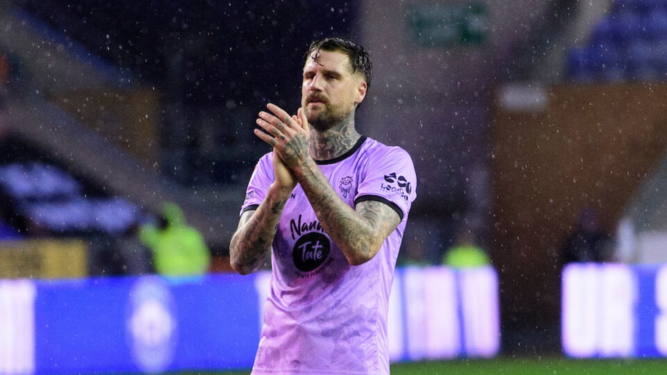A football player wearing a violet jersey applauds with a focused expression. Rain falls on the field, and blurred colorful lights illuminate the background.