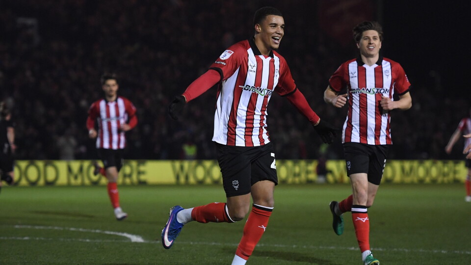 Football player in red and white striped jersey celebrates a goal on a floodlit field, with two teammates in the background. 