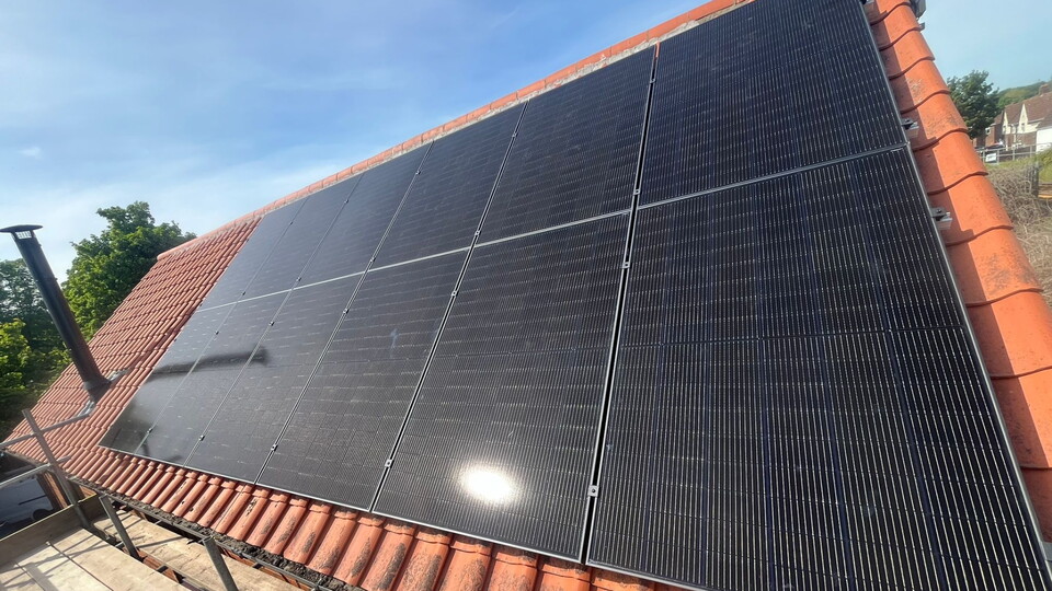 Solar panels on red-tiled roof under clear blue sky; panels are reflective and sunlit, with greenery in the background. The scene conveys sustainability.