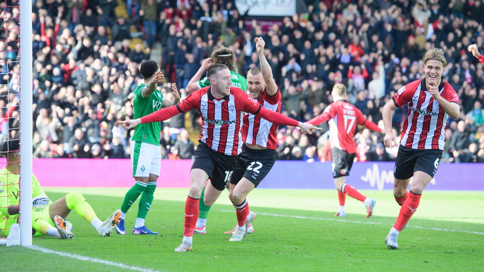 Soccer players in red and white stripes celebrating a goal near the goalpost as opponents in green stand nearby.