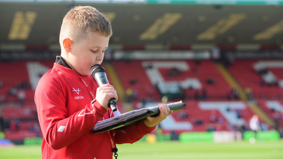 Child in a red jacket reads from a binder and speaks into a microphone on a football pitch with stadium seating in the background. 