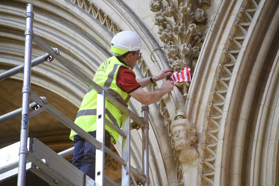 A man in a hi-vis yellow jacket attaches a red and white shirt to the iconic Lincoln Imp statue