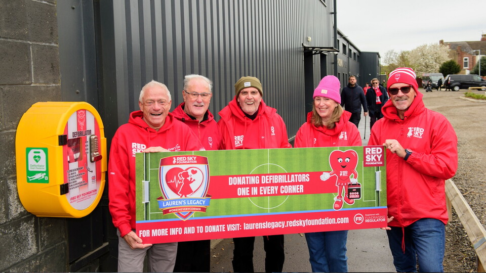 A group of five smiling individuals in red hoodies hold a banner promoting defibrillator donations. A defibrillator is mounted on a wall beside them.