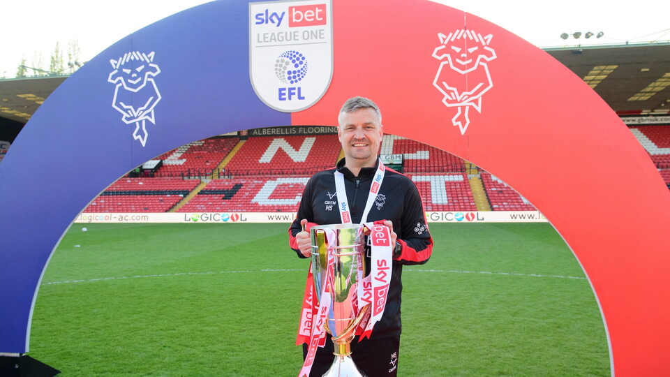 A smiling man holds the Sky Bet League One trophy on a football pitch. He stands under an arch bearing the league's logo. The mood is celebratory.